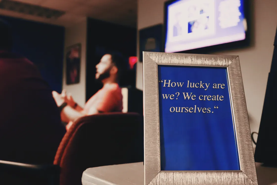 A framed card reading "How lucky are we? We create ourselves", me out of focus behind it, mid-talk.