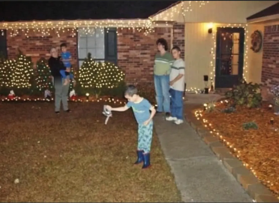 A brick house strung with Christmas lights after dark, a boy in pyjamas and rainboots scattering reindeer food across the lawn while the family watches from the path.